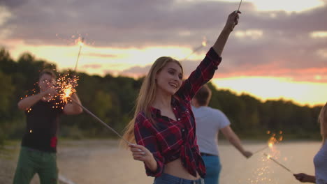 The-girl-in-red-plaid-shirt-and-denim-shorts-is-dancing-with-big-bengal-lights-in-her-hands-on-the-sand-beach-with-her-friends.-This-is-beautiful-summer-evening-on-the-open-air-party.